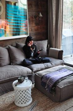 a woman sitting on top of a gray couch in front of a window next to a footstool