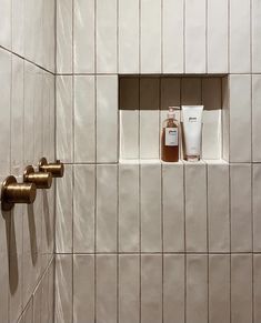 a white tiled shower with two gold faucets and a tube of lotion