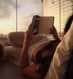 a woman reading a book while sitting on a couch in front of a large window