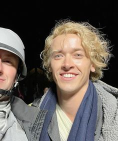 two men are posing for the camera with one man wearing a helmet and scarf around his neck