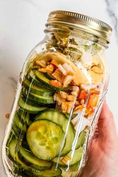 a person holding a mason jar filled with cucumber slices and other food items