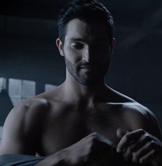 a shirtless man standing in the dark with his arm wrapped around his chest and looking off into the distance