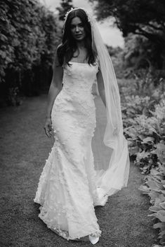 a woman in a wedding dress and veil walking down a path with flowers behind her