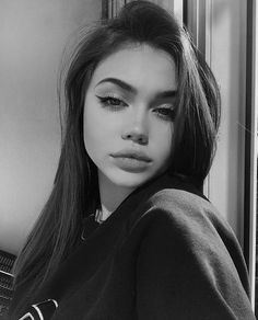 a black and white photo of a woman with long hair looking at the camera while leaning against a wall