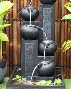 a water fountain made out of black rocks and some plants in front of a bamboo fence