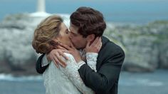 a man and woman kissing each other in front of the ocean with a lighthouse in the background