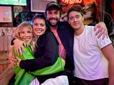 three people posing for the camera in front of a neon sign at a bar with their arms around each other