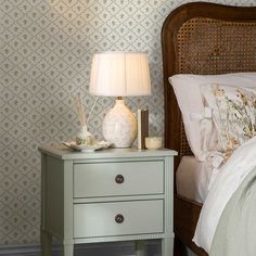 a nightstand with a lamp on top of it next to a bed and wall paper