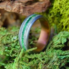 a wedding ring with green and black stripes on it sitting on mossy rocks in the woods