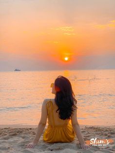 a woman sitting on top of a sandy beach next to the ocean under a sunset