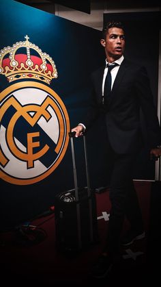 a man in a suit and tie holding a luggage bag next to a wall with the real madrid crest on it