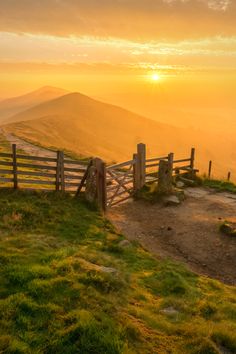 the sun is setting behind a wooden fence on top of a grassy hill with mountains in the background