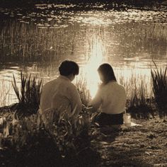 two people sitting on the ground next to a body of water with grass in front of them