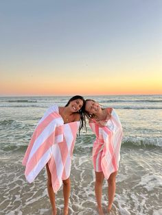 two women in pink and white towels standing on the beach with their arms around each other