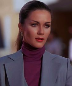 a woman wearing a gray suit and purple turtle neck top with her hair in a pony tail