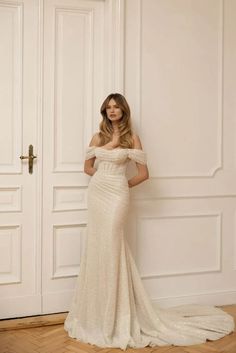 a woman standing in front of a door wearing a wedding dress with off the shoulder sleeves