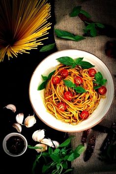 a white bowl filled with spaghetti and tomatoes next to garlic, pepperoni and basil