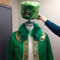 a green jacket with gold trim and fur collar being held up to it's head