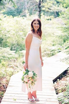 a woman in a white dress holding a bouquet on a wooden walkway surrounded by trees