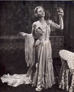 an old fashion photo of a woman holding a wine glass in her hand while standing next to a chair