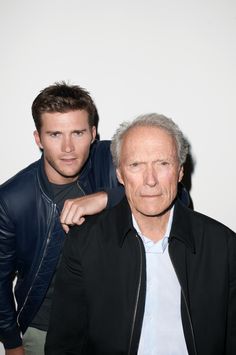 an older man standing next to a younger man in front of a white wall and wearing a black leather jacket