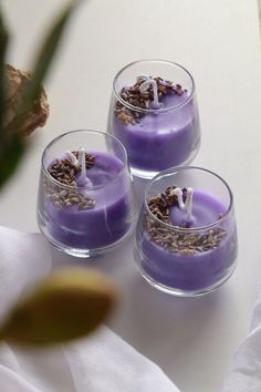 three small candles with lavender on them are sitting in glass cups next to a plant