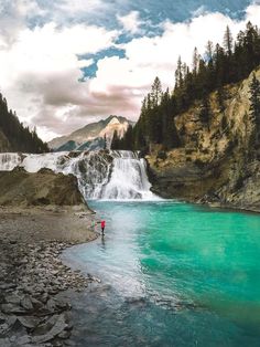 a person standing in the middle of a river next to a mountain with a waterfall