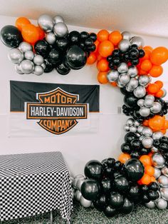 an orange, black and silver balloon arch with harley davidson logo on the wall next to a checkered table cloth