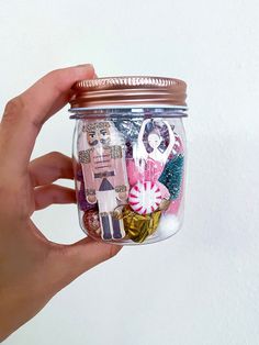 a hand holding a glass jar filled with magnets and paper art work on it