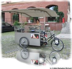 a woman is standing behind a food cart with a bicycle parked next to it in front of a brick building