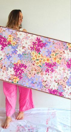 a woman in pink pants holding up a large floral piece of art on a bed