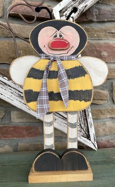 a wooden bee with a plaid bow on it's head and legs, standing in front of a brick wall