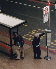 two people standing next to a bus stop on the side of the road with their hands in the air