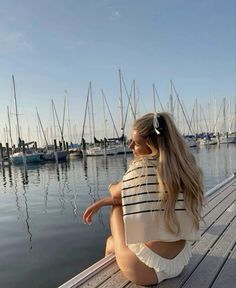 a woman sitting on top of a wooden pier next to water filled with boats in the background