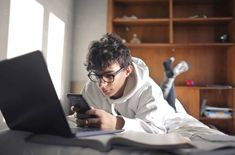 a young man laying in bed looking at his cell phone while using a laptop computer