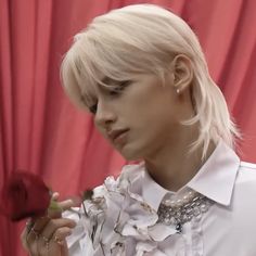 a woman with blonde hair holding a rose in front of a red curtain and wearing a white shirt