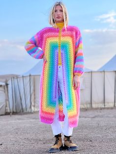 a woman standing in front of a fence wearing a rainbow colored sweater and white pants