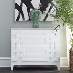 a white dresser with a green vase on top of it next to a large painting