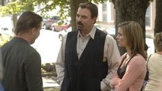 three people standing around talking to each other in the street with trees and buildings behind them