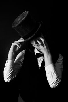 a man in a vest and hat covering his face with one hand while standing against a black background