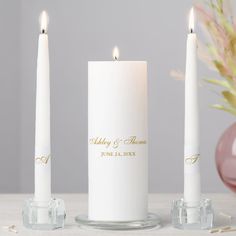 three white candles with gold lettering on them sitting next to a pink vase and glass candle holder