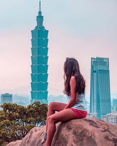 a woman sitting on top of a large rock in front of a cityscape