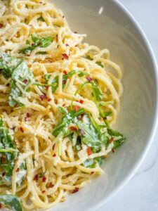 a white bowl filled with pasta and greens on top of a table next to a fork