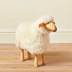 a white sheep standing on top of a hard wood floor next to a beige wall