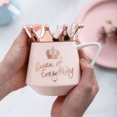 a person holding a pink coffee mug with gold crowns on it and the words queen of everything