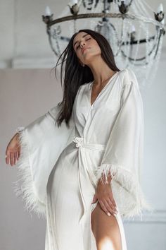 a woman wearing a white robe with feathers on the side and one leg up, standing in front of a chandelier