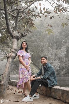 a man sitting next to a woman on a stone wall near a tree and water