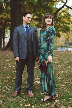 a man and woman standing next to each other in the grass with trees behind them