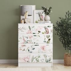 a white dresser with flowers painted on the front and sides, next to a potted plant