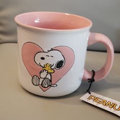 a pink and white coffee mug with a cartoon character on the side, sitting next to a tag that says peanuts